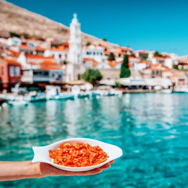 Elle tutulan kırmızı karidesli tabak. Arka planda balıkçı tekneleri ve Halki adasının limanı