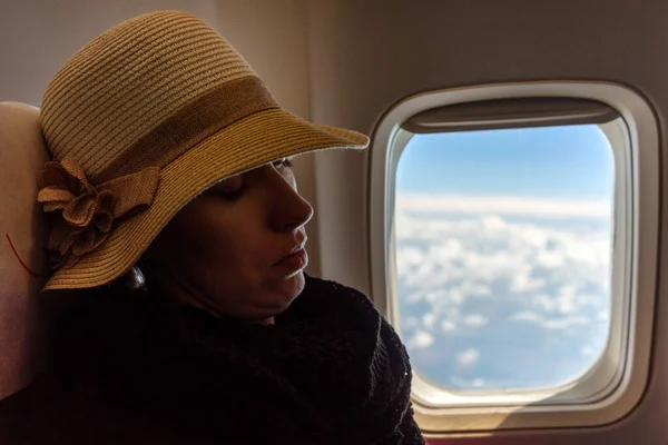 Woman with hat sleeping in airplane. Clouds on the sky in window of plane at background