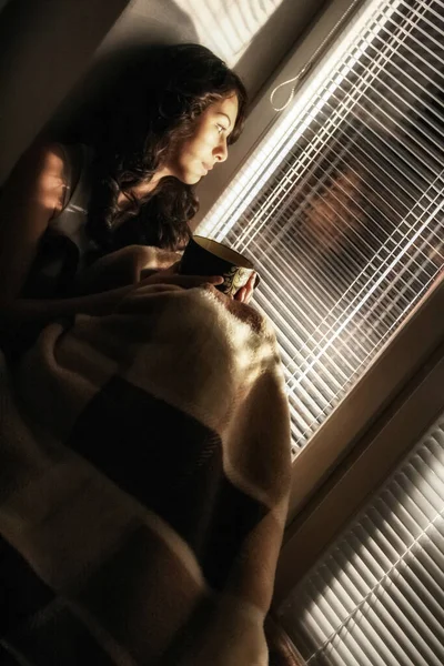 Sad woman sitting under blanket and looking outside room through window