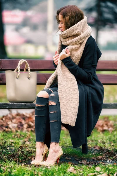 Fashionable dressed woman large scarf sitting lonely on bench in park.