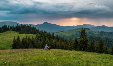 Çayırda oturan ve Slovakya 'daki Freat Fatra dağlarında renkli günbatımında bulutlarla turuncu gökyüzüne bakan bir adam.