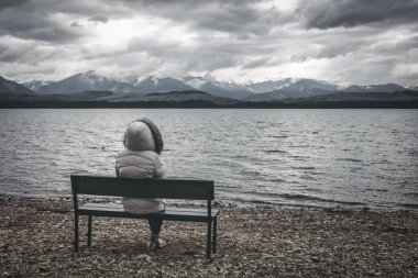 Göl kıyısında bankta oturan ceketli kadın. Karanlık bulutlu gökyüzü ve arka planda dağlar