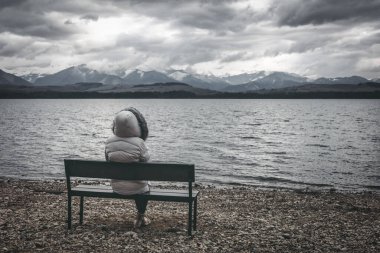 Woman in jacket sitting on bench on the shore of the lake. Dark Cloudy sky with mountains at background clipart