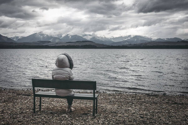 Woman in jacket sitting on bench on the shore of the lake. Dark Cloudy sky with mountains at background