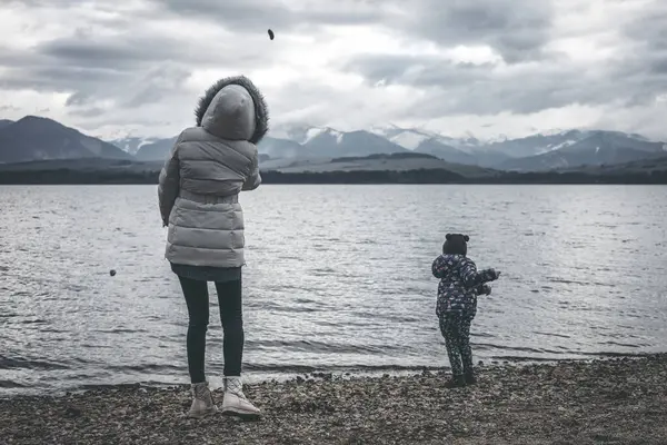 mother and daughter are throwing stones into the water on the shore of the lake. Dark cloudy sky and mountains at background