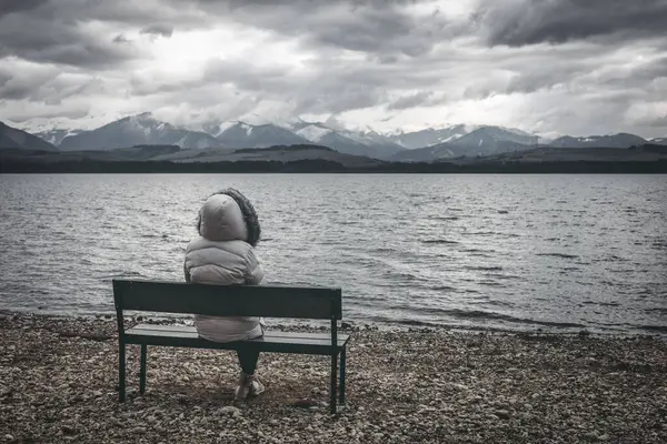 Žena Bundě Sedí Lavičce Břehu Jezera Tmavá Oblačná Obloha Horami Stock Snímky