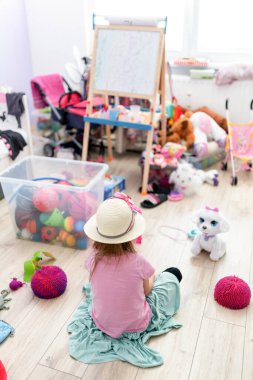 Kız odasında yerde oturup oyuncaklarla oynuyor. Çocuk odasında karışıklık var..