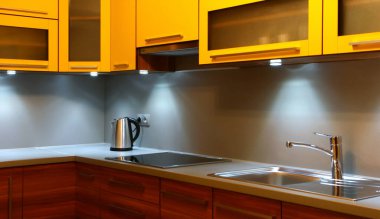 Modern mutfak içi sarı dolaplar, ahşap çekmeceler, paslanmaz çelik lavabo ve tezgahta elektrikli su ısıtıcısı, kabin ışıkları altında aydınlatılıyor.