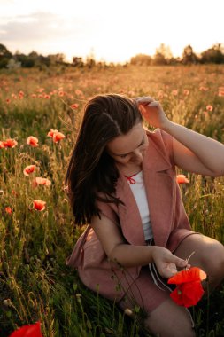Gün batımında haşhaş tarlasında kahverengi doğal saçlı ve tozlu gül takım elbiseli genç bir kadın. Polonya 'da yaz, Varşova.