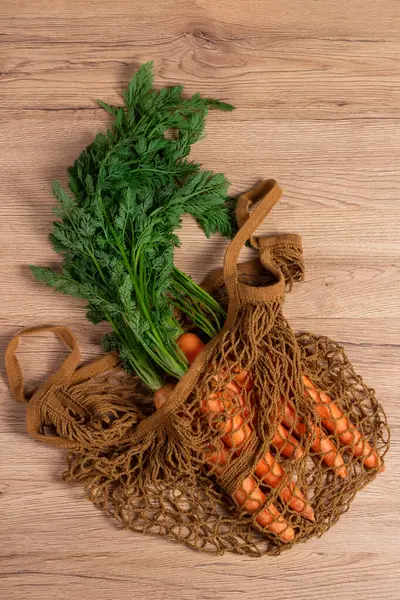 Tahta arka planda yeniden kullanılabilir ağ torbasında yeşil yapraklı taze havuç sürüsü. Net poşetteki havuç, çevre alışverişini, eko yaşam tarzını ve bitkisel gıda görsellerini betimliyor.