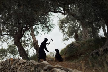 Zeytin Ağaçları Altında İnsan ve Köpek. Orak tutan bir adamın siluetli figürü. Yanında oturan siyah bir evcil hayvan var. Her ikisi de doğal bir manzarada zeytin ağaçlarının arka planına kurulmuş.