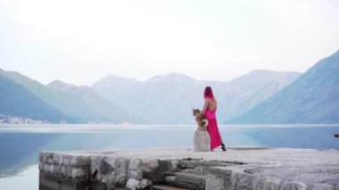 Pembe saçlı ve köpekli bir kadın sakin bir gölün kenarında, arka planda dağlar olan bir anı paylaşıyor. Bu sahne doğada huzurlu bir bağ yakalar.