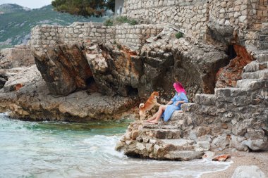 Pembe saçlı bir kadın ve bir Labrador Retriever antik deniz basamaklarında sessiz bir anı paylaşıyor, okyanus onların yanında fısıldıyor.