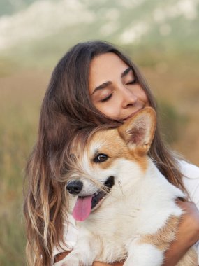 Neşeli genç bir kadın kırsal alanda gülümseyen bir Galli Corgi köpeğine sarılıyor. Paylaştıkları sevinç, açık arazinin dingin arka planına karşı hissedilir.
