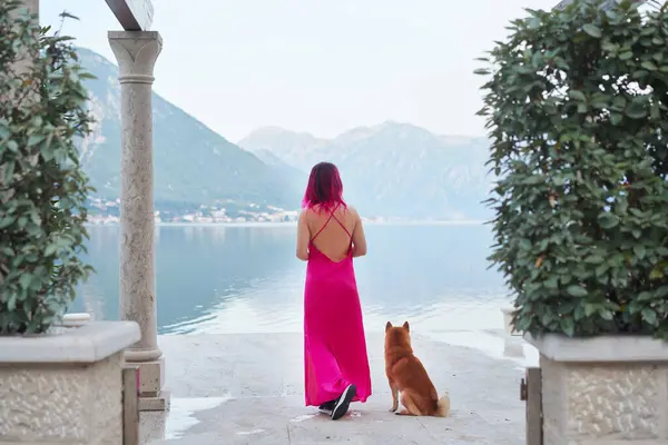 stock image A woman with pink hair in a flowing dress stands facing a breathtaking lakeside view, with a Shiba Inu looking towards her