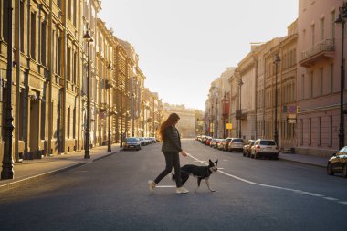 Gün doğumunda bir kadın, sınırdaki köpeğiyle karşıdan karşıya geçiyor. Binaları aydınlatan yumuşak bir ışık var. Görüntü, sabahın erken saatlerinde huzurlu bir anı yakalıyor..