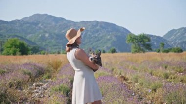 Bir kişi lavanta tarlasında duruyor, elinde küçük bir köpekle mor çiçek tarlalarına bakıyor. Uzak dağlar manzaranın güzelliğini ve huzurunu artırır..