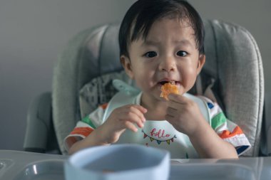 Sevimli, neşeli ve mutlu 1 yaşında Çinli bir çocuk bebek koltuğunda otururken yemek yiyor.