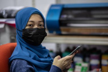 Toz ve enfeksiyonu önlemek için tıbbi maske takan Asyalı Müslüman kadın ofis saatlerinde akıllı telefon kullanıyor.