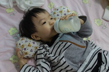 Asyalı Çinli bebek elinde süt şişesiyle yatağa uzanmış süt içiyor.