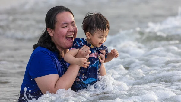 Asian Chinese mother with Happy 1 to 2 years old child enjoying playing on beach with splashing warm sea water