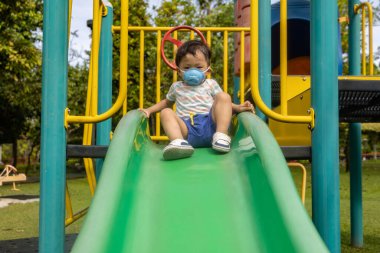 Parkta gündüz vakti tıbbi maskeyle oynayan Asyalı çocuk.