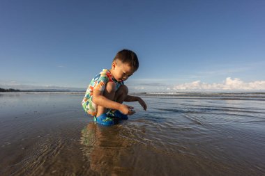 Neşeli bir çocuk, güneşli bir sahilde kumsalda oynar. Yazın kaygısız ruhunu ve açık hava aktivitelerinin neşesini somutlaştırır.