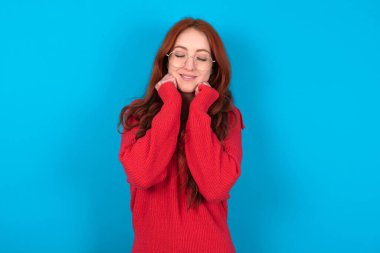 Mavi arka plan üzerine kırmızı kazak giyen genç bir kadın neşeyle sırıtıyor, hoş bir şey hayal ediyor, fotokopi odası. Hoş duygular konsepti.