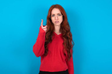Mavi arka planda kırmızı süveter giyen genç bir kadın hayal kırıklığına uğramış ve ön tarafı işaret ediyor.