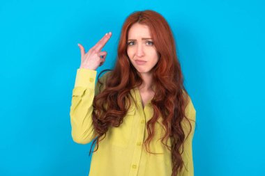 Mutsuz, kızıl saçlı, yeşil tişörtlü, mavi stüdyo arkaplanlı bir kadın intihar jesti yapıyor ve tabancayı eliyle taklit ediyor, dudaklarını kıvırıyor, iki parmağını şakakta tutuyor.