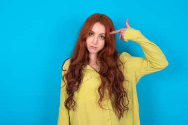 Mutsuz, kızıl saçlı, yeşil tişörtlü, mavi stüdyo arkaplanlı bir kadın intihar jesti yapıyor ve tabancayı elle taklit ediyor, kıvrımlı dudaklar iki parmağını şakakta tutuyor, ateş ediyor, her şeyden bıkıyor.,