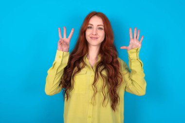 Mavi arka planda yeşil tişörtlü, beyaz saçlı genç bir kadın sekiz numaralı parmaklarıyla gülümseyerek kendinden emin ve mutlu bir şekilde onu işaret ediyor..