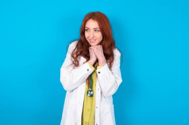 Mavi arka plandaki mutlu, genç, beyaz saçlı doktor kadın harika bir şey olacağını tahmin ediyor, mutlu bir şekilde yan yana bakıyor, ellerini yan yana tutuyor, mutlu bir ifadeye sahip..