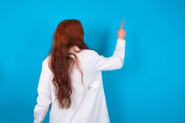 Mavi arkaplan üzerinde genç kızıl saçlı doktor kadın fotokopi alanındaki objeyi işaret ediyor. Arkanı dön.