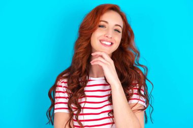 İyimser genç, beyaz saçlı, mavi arka planda çizgili tişört giyen kadın ellerini kısmen çapraz tutuyor ve ellerini çenenin altında tutuyor, zevkle kameraya bakıyor..