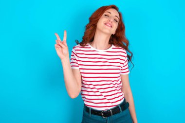 Mavi arka planda çizgili tişört giyen beyaz saçlı genç bir kadın. Kameraya bakıp zafer işareti yaparken gülümsüyor. İki numara..