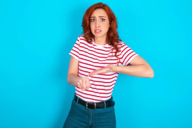 Mavi arka planda çizgili tişört giyen beyaz saçlı genç bir kadın. Aceleyle saati, sabırsızlığı, gecikmeye kızmış ve kızgın görünüyor..
