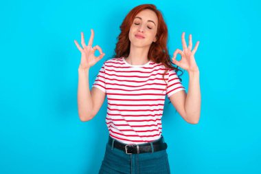 Mavi arka planda çizgili tişört giyen beyaz saçlı genç kadın rahatla ve gözleri kapalı parmaklarıyla meditasyon hareketi yaparken gülümse. Yoga konsepti.