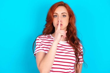 Beyaz saçlı, mavi arka planda çizgili tişört giyen genç bir kadın sessiz hareketler yapıyor, parmaklarını dudaklarının üzerinde tutuyor. Sessizlik ve gizli konsept.