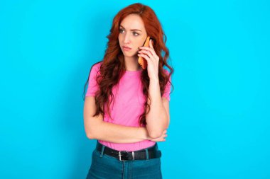 Üzgün, genç, beyaz saçlı, pembe tişörtlü, mavi arka planda, akıllı telefondan konuşan bir kadın. İletişim konsepti.