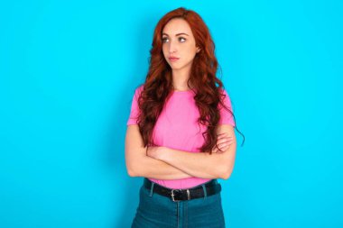 Mavi arka planda pembe tişört giyen, düşünceli, genç, beyaz, düşünceli bir kadın kollarını kavuşturmuş, düşünceli bir ifadeyle ne yapacağını düşünüyor.