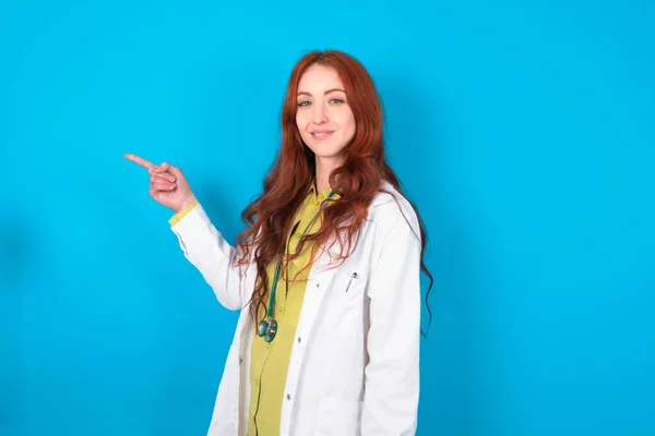 Profile photo of Young redhead caucasian doctor woman over blue background indicate finger empty space offer