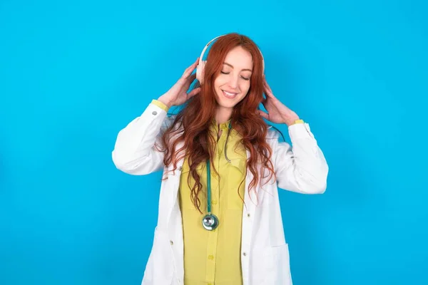 Young redhead caucasian doctor woman over blue background listen music on earphones
