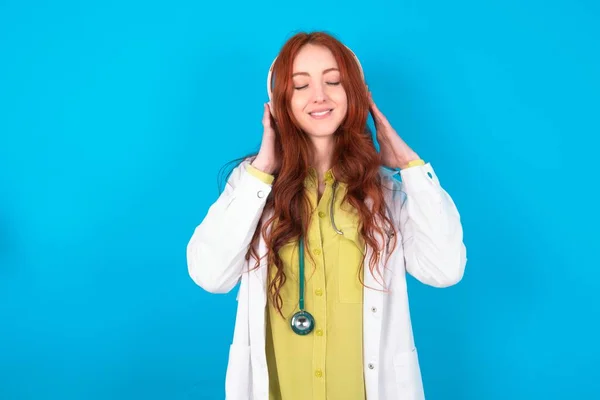 Young redhead caucasian doctor woman over blue background with headphones on her head, listens to music, enjoying favourite song with closed eyes, holding hands on headset.