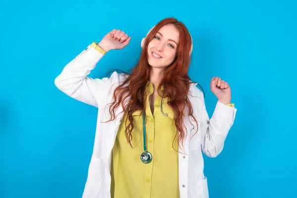 Carefree Young redhead caucasian doctor woman over blue background with toothy smile raises arms dances carefree moves with rhythm of music listens music from playlist via headphones