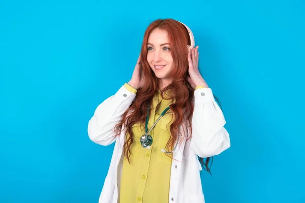Young redhead caucasian doctor woman over blue background wears stereo headphones listens music concentrated aside. People hobby lifestyle concept