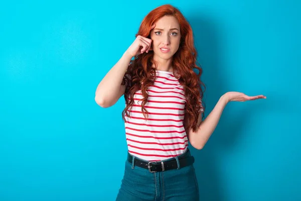 Young caucasian redhead woman wearing striped t-shirt over blue background confused and annoyed with open palm showing copy space and pointing finger to forehead. Think about it.