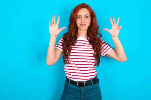 stock image Young caucasian redhead woman wearing striped t-shirt over blue background showing and pointing up with fingers number nine while smiling confident and happy.