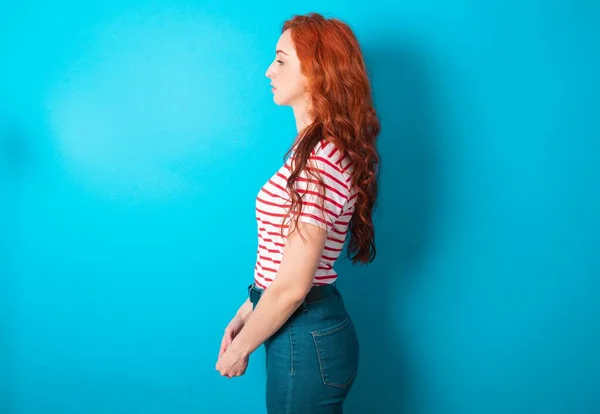Young caucasian redhead woman wearing striped t-shirt over blue background looking to side, relax profile pose with natural face with confident smile.