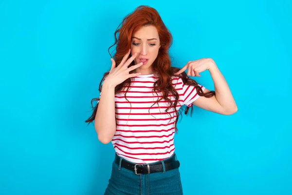 stock image Shocked Young caucasian redhead woman wearing striped t-shirt over blue background  look surprisedly down, indicates at blank  with fore finger, Scared model